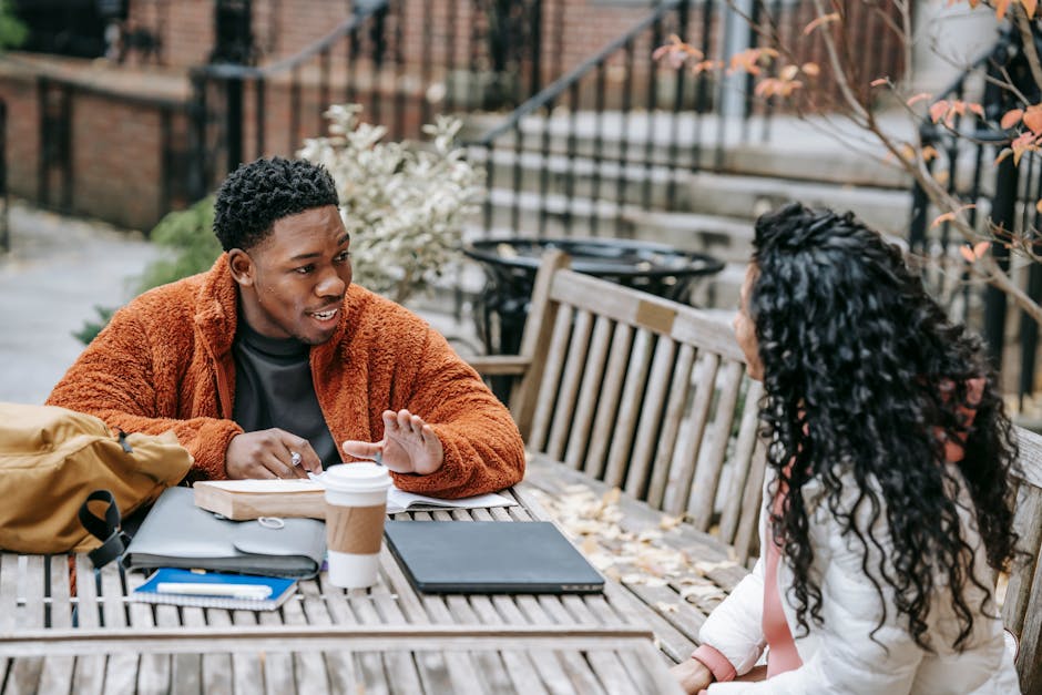 Two young adults discussing a project outdoors with coffee and laptops.